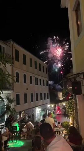 The video shows a narrow street at night, possibly in a European town, with a building on the left and a lighter colored building on the right. There is a fireworks display occurring in the sky above the street, with bursts of pink, purple, and green. People are gathered in the street, sitting at tables outside what appears to be a restaurant or bar. Some individuals are standing and looking up at the fireworks. There are several cars driving down the street, their headlights and taillights visible. The overall atmosphere appears festive.