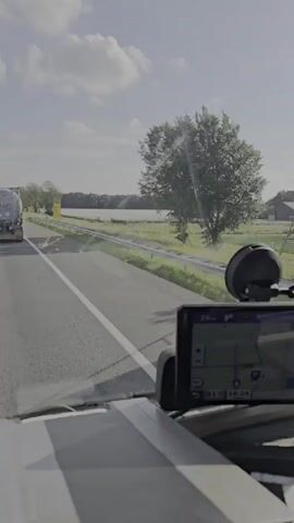 The video shows a view from the driver's seat of a vehicle, looking out the windshield. A GPS device is mounted on the dashboard in the foreground, displaying a map. The vehicle is on a highway with a white lane marker. In the distance, a truck with a white trailer is visible in the left lane. To the right of the road, there is a guardrail, grassy fields, trees, and a body of water. The sky is partly cloudy, and the sun is shining. The video appears to be recorded during the daytime.
