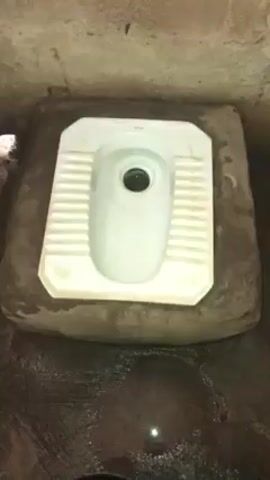 The video shows an unflushed squat toilet in a cement room. The toilet bowl is white with a dark stain in the center. The surrounding cement is damp and discolored. The walls of the room are rough and appear unpainted.
