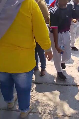 The video shows a group of people, including adults and children, standing outdoors in what appears to be a crowded area. The lighting suggests it is daytime. One person in the foreground wears a bright yellow long-sleeved shirt and blue jeans. Another person, also in the foreground and slightly to the right, wears a black t-shirt with white stripes on the sleeves and white pants. Several children are visible, some with painted faces. One child is holding a small white cup. In the background, more people are gathered, and there are some buildings and trees. The ground is paved and shows some cracks. The overall impression is of a gathering or event taking place in a public outdoor space.
