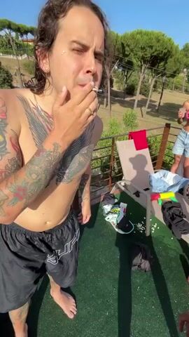 The video opens with a close-up of a man smoking a cigarette, his arm covered in tattoos. The camera pans out to reveal a group of people by a swimming pool. Two men and two women are visible. One woman is sitting on the edge of the pool with her legs in the water. Another woman is in the pool, swimming. A man is floating on a yellow inflatable ring. The background shows trees and a clear blue sky. The lighting suggests it is a sunny day.