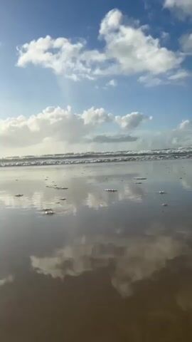 This video is a first-person perspective shot of a beach. The camera is low to the ground, looking out towards the ocean. The sky is blue with scattered white clouds, some of which are reflected in the wet sand. Gentle waves are breaking on the shore in the distance. There are small, foamy bubbles or detritus scattered on the wet sand. The lighting suggests it is daytime, possibly late afternoon or early morning, with the sun casting rays through the clouds. No people or other objects are visible. The overall atmosphere is calm and serene.