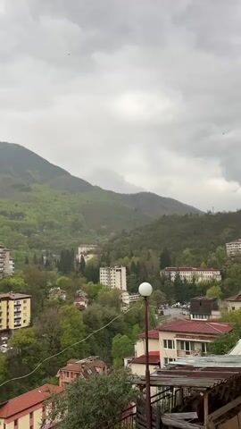 This video captures a panoramic view of a mountain town or resort area on an overcast day. The camera slowly pans from right to left, revealing a landscape dominated by lush green, forested mountains that rise steeply in the background. Scattered throughout the hillsides are numerous multi-story buildings, appearing to be apartment blocks or hotels, many with balconies and varying architectural styles, often featuring white, yellow, or pink facades with red roofs. Some larger structures with multiple windows, possibly commercial or public buildings, are visible further down in the valley.

The foreground includes rooftops of closer buildings and a street lamp. Trees and dense foliage are abundant, indicating a verdant environment, likely in spring or summer. A road can be glimpsed winding through the valley. The sky is largely covered with thick, gray clouds, creating a soft, diffused light, and some distant mountains appear hazy, possibly due to mist or rain. No people or specific activities are clearly visible, and there is no legible text on any of the structures. The scene conveys a sense of quiet and tranquility amidst nature.