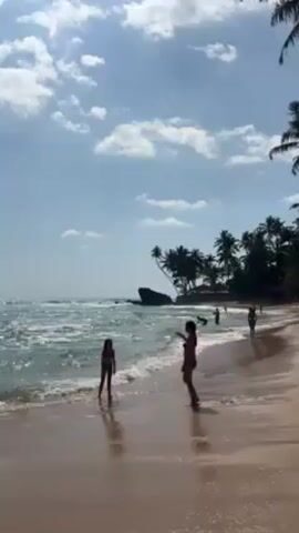 The video shows a tropical beach with several people. In the foreground, a woman and a child stand near the water's edge. The woman is wearing a bikini. The child wears a swimsuit. They are walking along the shoreline as waves break. In the background, other individuals can be seen further down the beach. The sky is partly cloudy with a blue hue. Palm trees line the edge of the beach. Rocks are visible in the ocean.