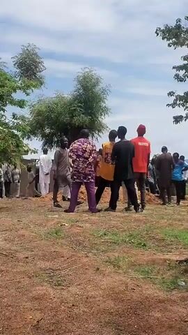 A panning outdoor shot reveals a group of people gathered under trees. Some are standing, and others are seated on chairs. The ground is dry and dusty with sparse vegetation. The sky is partly cloudy.