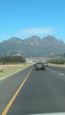 The video shows a road trip along a highway. Multiple cars are traveling in the same direction. The road is asphalt and is bordered by a yellow line. Beyond the road, there is a grassy field with a fence. In the distance, a large, rocky mountain range stands against a clear blue sky.