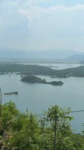 This is a vertical, handheld, medium shot video looking out over a large body of water. The water is a calm, deep blue-green and reflects the sky. Several green, tree-covered islands dot the lake. In the background, rolling, green hills and mountains fade into a hazy blue distance under a bright, partly cloudy sky. In the foreground, the tops of leafy green trees and a thin, dark wire are visible. The scene appears to be a natural landscape, possibly a reservoir or a large lake in a mountainous region. There are no people or discernible human activity visible in the shot, except for the wire which could be a power line.