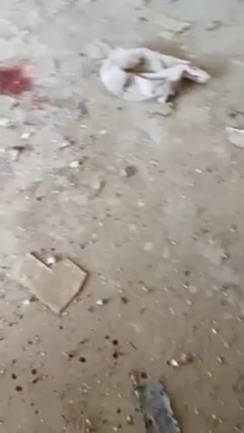 The video shows a room with a concrete floor that is covered in debris and what appear to be splatters of blood. The camera pans across the room, revealing discarded shoes, metal framing, and other miscellaneous items. A man wearing a gray outfit and a head covering walks into the doorway and looks outside. There are other people visible outside the building. The overall impression is that the room is in disarray and may be the site of a violent event.