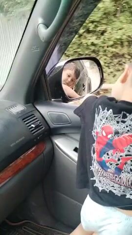 This video shows two young children, likely brothers, inside a car. One child is in the foreground, facing away from the camera and looking out the side window. They are wearing a black t-shirt with a prominent Spider-Man graphic. The other child is visible in the car's side mirror, looking back at the child in the foreground. The scene appears to be set in a residential area, with a fence and greenery visible outside the car window. The time of day appears to be daytime, with natural light illuminating the scene. The overall mood is playful and innocent, as the children seem to be interacting with each other. There is no visible text in the video, other than the "Spider-Man" logo on the t-shirt.