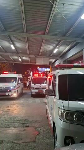 The video shows an outdoor scene, likely a covered driveway or entrance, at night. Three white ambulances with red and blue flashing lights are visible. The ambulance in the foreground is partially obscured, but its side and front are visible. The second ambulance is in the middle ground, and the third is further back and to the left. Several people are seated or standing near the left side of the frame, appearing to be waiting or observing. The setting appears to be a medical facility, possibly a hospital, given the presence of multiple ambulances and the overall ambiance of a nighttime emergency response. The lighting suggests it is dark outside, with artificial lights from the covered area and the ambulances illuminating the scene. There is no indication of weather conditions. The overall mood is one of urgency and preparedness. Visible text includes "AMBULANCE" on the front of the ambulance in the middle ground and "EMERGENCY" on the side of a vehicle further in the background. The location context of Banda Aceh, Indonesia, places this scene within that geographical region.