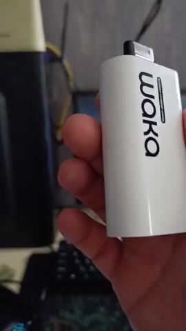 A person's hand is holding a white disposable vape device with the word "waka" printed vertically in black text. A barcode is also visible next to the word "waka." The background is blurry, but appears to be an indoor setting with a desk and computer equipment.