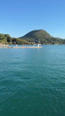 A vertical, stabilized, first-person perspective video pans across a calm, turquoise bay under a clear blue sky. In the foreground, the rippling water fills most of the frame. On the left, a wooden pier extends into the water, marked by a red and white pole. In the mid-ground, several sailboats and a larger motor yacht with an orange stripe are moored. The background features a lush green, tree-covered hill with a peak, and a series of smaller hills receding into the distance. The video appears to be filmed from a boat or the edge of a dock.