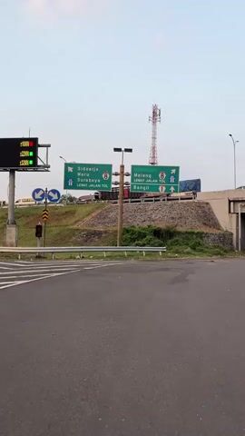 This is a vertical, medium-angle shot of a road intersection with highway signs. In the foreground, there is asphalt paving, and a white line indicates the edge of a lane. A metal guardrail runs along the side of the road. Behind the guardrail, a grassy embankment rises, topped with a stone retaining wall. On top of the embankment, two large green highway signs are mounted on a single pole. The sign on the left indicates directions to Sidoarjo, Waru, and Surabaya via Jalan Tol 8. The sign on the right indicates directions to Malang via Jalan Tol 8 and Bangil via Jalan Tol 1. Several trucks and cars are visible on the elevated highway behind the signs. A tall red and white communication tower is visible in the background, to the right of the signs. The sky is overcast.