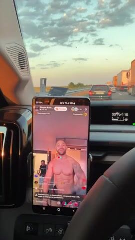 The video begins with a close-up of a smartphone mounted on a car's dashboard. The phone screen displays a live stream featuring a shirtless, muscular man with tattoos on his arms and chest, posing with his arms raised and mouth open. The background of the live stream is dimly lit with red and blue lights. The phone's interface shows comments and likes. Outside the car, through the windshield, a traffic jam is visible on a highway during sunset. The sky is filled with clouds illuminated by the warm orange and pink hues of the setting sun. Numerous cars and large trucks are stationary on the road, indicating a standstill. The car's dashboard displays various indicators, including speed and temperature. The video then pans slightly to the left, showing more of the traffic and the highway environment. The focus shifts between the phone screen and the road ahead. The camera is positioned inside the car, capturing the view from the driver's perspective.