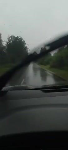 A first-person view from inside a vehicle driving down a wet road during a rain shower. The windshield is covered in raindrops, and the windshield wipers are intermittently clearing the view. The road is asphalt and appears to be a rural highway lined with trees on both sides. The sky is overcast and gray, contributing to the gloomy atmosphere. The car is moving forward, indicated by the motion blur and the appearance of the road stretching ahead.