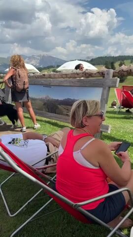The video shows a group of people relaxing and conversing outdoors on a sunny day. In the foreground, a man in a white t-shirt and khaki shorts lies in a red lounge chair, facing away from the camera, with his eyes closed. Beside him, a woman in a bright pink tank top and dark shorts holds a smartphone in her right hand. She is looking to her left, and her mouth is open as if she is speaking or laughing. Behind them, several people are standing near a wooden display board with an image of a mountainous landscape. A woman with curly blonde hair and a backpack stands facing the display, while a man in a red t-shirt and blue shorts stands next to her, also looking at the display. A black dog stands between them. Further in the background, a few people are seated in lounge chairs under white umbrellas, and a range of mountains can be seen under a partly cloudy sky. The environment appears to be a grassy outdoor area, possibly a park or a scenic viewpoint.