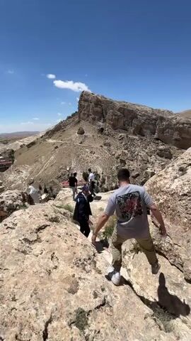 Several people are navigating a rocky, mountainous landscape on a sunny day. The sky is blue with a few scattered clouds. The terrain is arid and rocky, with sparse vegetation. In the background, there is a large, distinctive rock formation and a village. The individuals in the video are walking down a rocky path. One person extends a hand to assist another. They appear to be tourists or explorers taking in the scenic view.