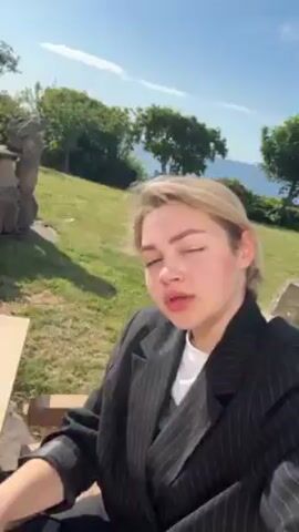 Here is a description of the video:

The video shows a woman sitting outside on a sunny day at a wooden table. She's wearing a black pinstripe blazer and dark pants. On the table are a glass with a green liquid, a carton of Alpro hazelnut milk, and a blue bowl. She picks up the glass and says, "I'm drinking my matcha." She pauses, then adds, "And this day is absolutely gorgeous." The background includes wooden chairs, green grass, trees, and a stone building or wall.