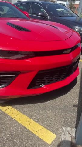 The video pans across the front and side of a bright red Chevrolet Camaro parked in a parking lot. The car has black rims and tinted windows. In the background, other cars and buildings are visible. The parking lot has yellow lines marking the parking spaces. The sun is shining, casting shadows.