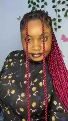 The video shows a woman standing indoors. She has dark hair that is styled into red-burgundy braids. She is wearing a long-sleeved black floral-print shirt. She is looking at the camera, then turns her head to the right. She then moves her hair from the front of her face. The background includes a white wall decorated with a green vine and a pink butterfly sticker.