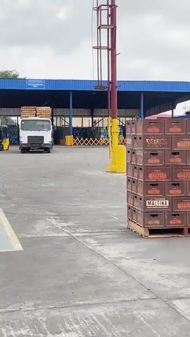 The video depicts an active outdoor industrial yard, likely a beverage production or distribution facility, under a cloudy sky. Large stacks of brown crates, prominently labeled "MALTINA," are visible on wooden pallets in the foreground.

At the 0:01 mark, a person is seen operating a yellow and black forklift, moving across the yard. The forklift appears to be transporting crates or pallets. Surrounding the area are numerous stacks of empty wooden pallets, indicating a high volume of goods movement. In the mid-ground and background, there are large piles of packaged goods, possibly beverages, wrapped in plastic and stacked on pallets.

The industrial setting is further emphasized by several buildings with blue and green roofs, and imposing gray industrial silos, some of which bear the label "SYMAGA." A white tank with the letters "PI" visible on its side can also be seen. The scene captures the busy operations of a logistics or manufacturing site in Sabon Gari Nassarawa, Nigeria.