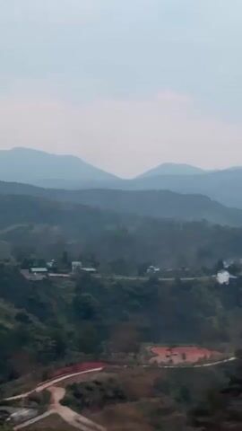 The video shows a scenic landscape of rolling hills and mountains under a cloudy sky. In the foreground, green vegetation and a dirt road are visible. Some buildings are scattered across the hillside. As the camera moves, trees come into view, partially obscuring the landscape. The overall atmosphere is tranquil and natural.