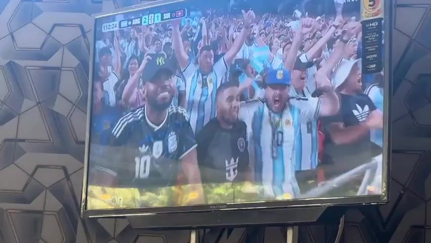 The video shows a television screen displaying a soccer match. The screen shows a crowd of jubilant fans wearing Argentina jerseys, cheering enthusiastically. The score on the screen indicates that Argentina is playing against Switzerland, with Argentina leading 2-1. The fans are close to the screen, some are waving flags and holding up their fists in celebration. The lighting is bright, suggesting it is daytime or the stadium lights are very strong. The TV is on a stand with a dark base, and a patterned wallpaper is visible in the background. The camera appears to be handheld and slightly shaky, as the video is not perfectly steady. There are reflections on the screen from the environment where the video is being recorded.