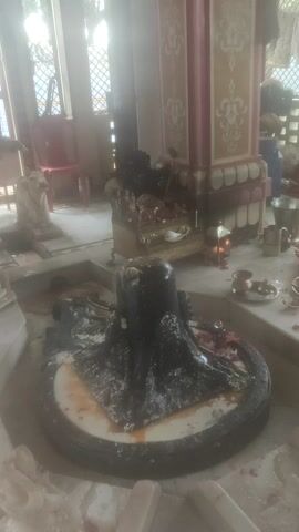 The video shows the interior of what appears to be a temple. In the foreground, a dark stone object, possibly a deity statue or altar, is partially covered in white powder and orange residue. Behind it, several people are present, some in yellow or orange clothing, engaged in activities that suggest a religious ceremony or offering. One person is leaning over the stone object, possibly performing a ritual. The background reveals architectural details, including patterned walls and a lattice-like window. The lighting is dim, with sunlight filtering through the window, creating a hazy atmosphere. The camera appears to be handheld and is tilted at an angle, with occasional shaky movements.