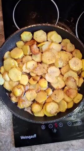 A top-down shot shows sliced potatoes being fried in a black frying pan on a black induction stove. The potatoes are golden brown and have been seasoned with salt and pepper. The stove has a digital display that shows the number 5. The scene is lit by overhead lighting.