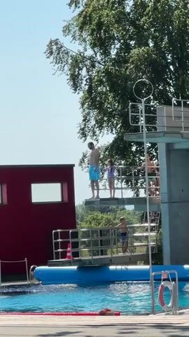 At 00:00, a man in blue swim trunks stands on a diving board with a red building to his left and trees behind him. At 00:01, the man jumps off the diving board. At 00:02, the man hits the water, creating a splash. At 00:03, the splash continues to expand as the man disappears underwater. At 00:04, the water continues to settle. Other people are visible on the diving platform and around the pool.