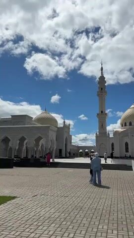 The video pans across a courtyard in front of a white mosque. The mosque features multiple domes and tall minarets. In the foreground, two people stand facing away from the camera, looking towards the mosque. Other people are visible in the distance, walking around the courtyard and entering the mosque. The sky is blue with white, fluffy clouds. The ground is paved with light-colored bricks. A fountain is visible in front of the main mosque building.