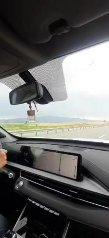 This is a first-person point-of-view video taken from inside a car, driving on a highway. The dashboard is visible in the foreground, with a large touchscreen display showing a navigation map. A hand is resting on the steering wheel. The windshield offers a view of the road ahead and the surrounding landscape. Several other vehicles, including cars and a van, are on the highway. The sky is overcast, and mountains are visible in the distance. The video is shot from a moving vehicle, indicating travel.