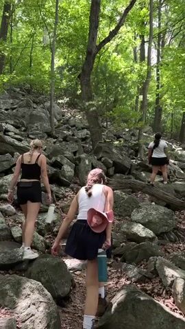 The media file shows four individuals hiking on a rocky, wooded trail. The subjects are three women in the foreground and one in the distance. They are dressed in casual, athletic wear suitable for hiking, including shorts, tank tops, and athletic shoes. Each person is carrying a water bottle. One woman has a pink hat and a blue water bottle. Another is wearing denim overalls. The trail is characterized by large, grey rocks and lush green trees, suggesting a natural setting in a forest. The sun is shining through the canopy, indicating it is daytime and the weather is likely pleasant. The path appears to be uphill, and the rocky terrain suggests a challenging but scenic hike. The overall mood seems to be one of enjoyment and physical activity in nature. No visible text is present in the media file. The location context is Edgemont, United States, which aligns with the appearance of the natural environment.