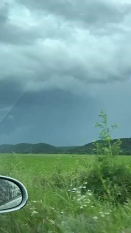 The video shows a view from inside a moving vehicle looking out at a field of tall green grass under a dark, cloudy sky. There are hills in the distance and telephone poles in the field. As the car continues moving, various plants and trees appear to be close to the road. The side mirror of the car is visible in the bottom left corner of the frame.