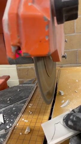 A close-up, low-angle shot shows an orange tile saw with a spinning blade cutting a small white tile. The tile has a black curved line drawn on it, indicating a cut line. A gloved hand is holding the tile against the saw's track. The background shows other broken tile pieces and a brick wall.