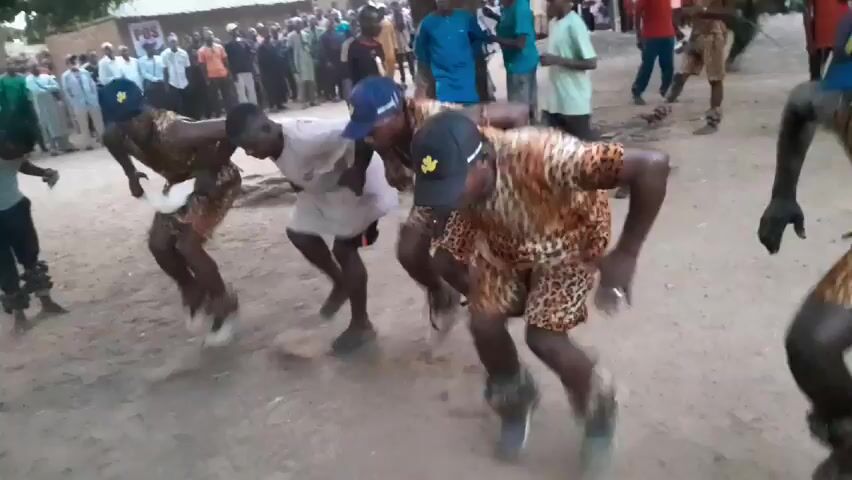 A group of men perform a traditional dance in a village setting. The dancers wear leopard-print clothing and baseball caps. Some of them have bells or noise-makers attached to their ankles. The dance involves rhythmic bending and swaying. Spectators watch from the background, creating a lively atmosphere. The setting seems to be a sandy ground in a village square with a tree in the background.
