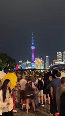 The video shows a nighttime view of the Shanghai skyline, dominated by the Oriental Pearl Tower illuminated in purple and blue lights. A crowd of people stands in the foreground, some holding umbrellas and phones. The buildings in the background are lit up, with the Shanghai Tower prominently featuring blue lights. The overall atmosphere appears to be one of a public gathering or viewing event.