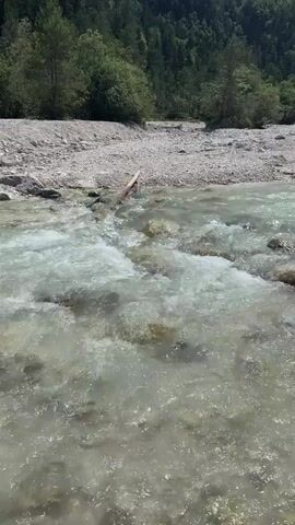 A clear, shallow river flows over rocks and pebbles in the foreground. A wooded mountain range with snow-capped peaks rises in the background under a bright blue sky. The riverbank is covered in small stones and a few scattered logs. The camera pans slightly upwards, revealing more of the mountainous landscape. There are no people or animals visible in the video. The weather appears to be sunny and warm.