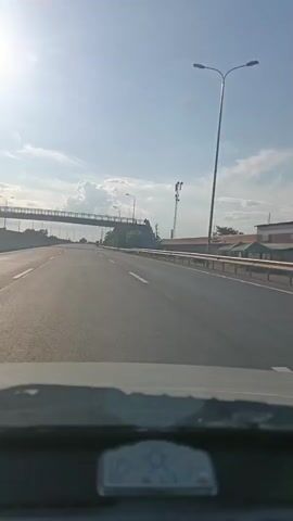 The video shows a view from inside a vehicle driving on a multi-lane highway on a sunny day with some clouds. A pedestrian overpass is visible on the left side of the highway, crossing over it. Further down the highway, a tall tower, possibly a cell tower, is visible on the right. Trees line the sides of the highway. The perspective is looking forward through the windshield.