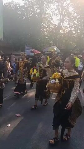 This video shows a parade or procession. Several young women, dressed in traditional, ornate costumes featuring black, gold, and silver, are walking in the street. They are all wearing elaborate headpieces with yellow flowers and have their hair pulled up. Each woman is carrying a woven basket filled with various fruits, including bananas, apples, and what appear to be coconuts or gourds, as well as some decorative items. The women are walking in formation, and some appear to be performing a synchronized dance or movement. Behind them, there are spectators lining the street, some sitting on the curb. In the background, there are trees, buildings, and a parade float with the number 09 on it. The lighting suggests it is daytime, possibly late afternoon, with the sun creating a bright, hazy effect. There are also some vendors or food stalls visible in the background. The overall atmosphere appears festive and cultural.