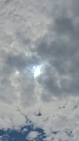A video shows a sun partially obscured by clouds. The sun appears as a bright, white orb with rays of light emanating from it. The clouds are a mix of white and gray, suggesting a partly cloudy day. The sky below the clouds is a clear blue. The sun is positioned in the center of the frame. The video is shot from a low angle, looking up at the sky. There are no people or objects visible in the frame besides the sun and clouds.