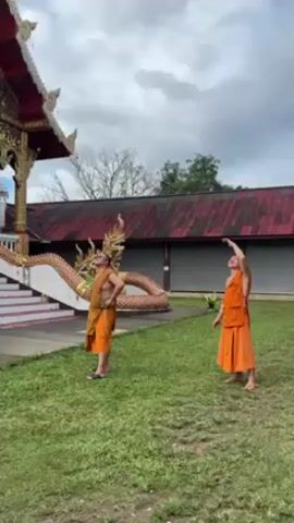 Two monks in traditional orange robes stand on a lawn next to a temple. They gaze upwards and point. Behind them is a dragon statue, temple steps, and a building with a red roof. The sky is cloudy.