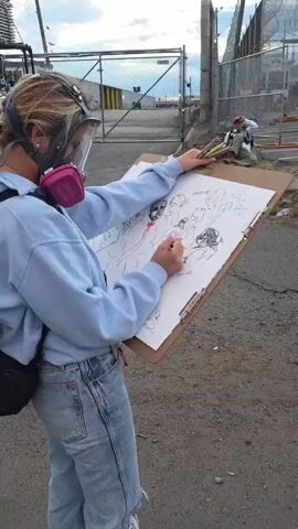 A woman wearing a face shield and pink respirator stands outdoors and draws on a clipboard. She is wearing a light blue sweatshirt and jeans, and has a black bag slung over her shoulder. In the background, a chain-link fence runs along the edge of the area, with other people standing around. The ground is dirt and gravel. The woman is actively drawing, using a pencil or pen to add details to her sketch. The drawing depicts several figures, seemingly of people. The sky is visible in the background, suggesting it's either early morning or late afternoon.