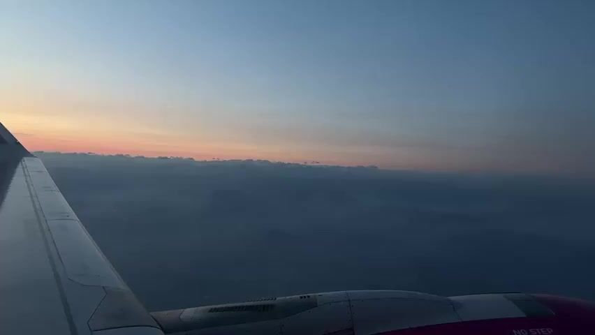 The video shows a view from an airplane window during sunrise. The left side of the frame is dominated by the wing of the aircraft, which is white and has a gray leading edge. The right side of the frame shows the engine nacelle, which is primarily purple. The sky transitions from a deep blue at the top to a pale orange and pink hue along the horizon, indicating sunrise. Below the horizon, there is a layer of clouds or mist, and beyond that, the silhouettes of mountains are visible, suggesting the airplane is flying over a mountainous region. The lighting conditions indicate early morning or late evening. The scene is serene and depicts a typical aerial view during a flight.