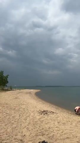 The video shows a sandy beach with a person in the distance sitting on the sand. The water is calm and blue, with land visible on the horizon. The sky is overcast with dark clouds. There is a sign on a pole in the distance, and a tree to the left of the frame. The video pans slightly to the left and then right.