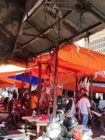The video shows an outdoor market, possibly in Southeast Asia. Several stalls are visible, covered with orange and blue tarps. Hanging from the stalls are cuts of raw meat. Several people are present, some standing near the meat, others sitting. There are also several parked motorbikes. The market appears to be open and active.