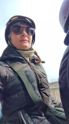 Here is a concise description of the video:

The video shows a woman riding on the back of a scooter on a mountainous road. The woman is wearing a black puffer jacket, sunglasses, and a yellow helmet. The man driving the scooter is wearing a purple helmet and a dark vest. The road is paved and there are metal guardrails on the side. The sky is overcast and the surrounding landscape appears to be mountainous with some trees.
