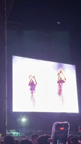 The video shows a live concert performance. A large screen displays three performers wearing sequined outfits. The central figure appears to be the lead singer, while the other two are backup vocalists or dancers. The camera zooms in on the screen, focusing on the backup vocalists and their expressions. They are singing and raising their hands, engaging with the audience. The stage lighting is bright, creating a vibrant atmosphere, and the crowd is visible in the foreground. A person in the crowd is holding up a phone to record the performance.