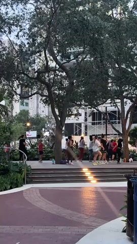 A group of about ten people are participating in an outdoor fitness class. They are gathered on a raised platform with tiered seating and are arranged in a line. The individuals are mostly women, and they are dressed in athletic wear. Some are performing lunges and other exercises, while others appear to be following instructions from a person at the front of the group, who may be an instructor. The setting is a park-like area with large trees providing shade. In the background, there are buildings and streetlights. The lighting suggests it is late afternoon or early evening.