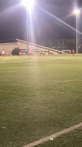 The video shows a group of people playing soccer on an artificial turf field at night. There are bright stadium lights illuminating the field. Several players wearing red and yellow jerseys are visible. Some players are running, while others appear to be kicking a soccer ball. The field has white lines marking boundaries and a goal net is visible in the background. Buildings and trees can be seen beyond the field.